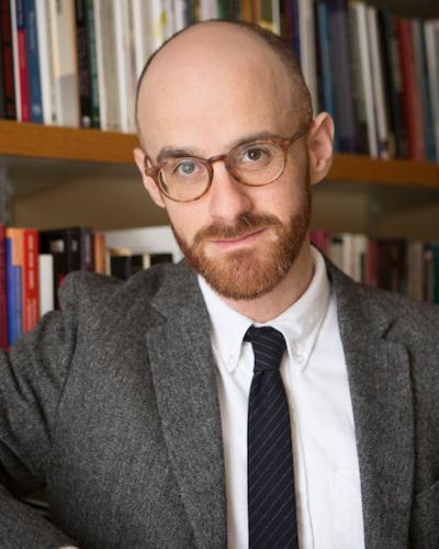 Alexander Livingston poses in front of a full bookshelf wearing orange-framed glasses